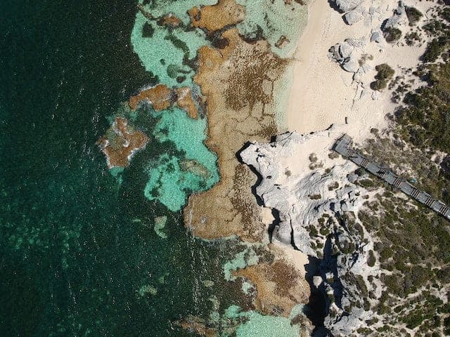 Aerial view looking straight down on a rocky coastline: deep teal water and turquoise shallows over submerged reef on one side, pale sand and white limestone outcrops on the other, with a timber boardwalk running down to the shore