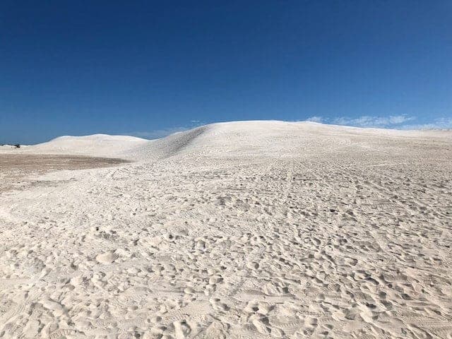 Broad white sand dunes rising against a deep blue sky, the sand marked with footprints and vehicle tracks