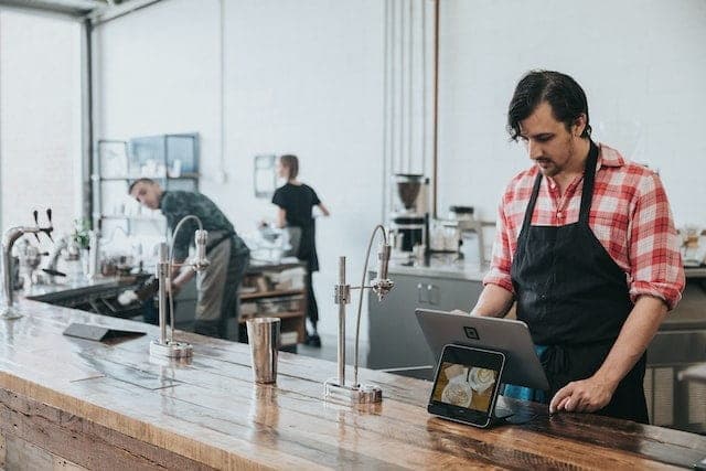 Barista in a check shirt and apron working at a long timber café counter with brewing equipment and a tablet, two colleagues behind him in the open kitchen