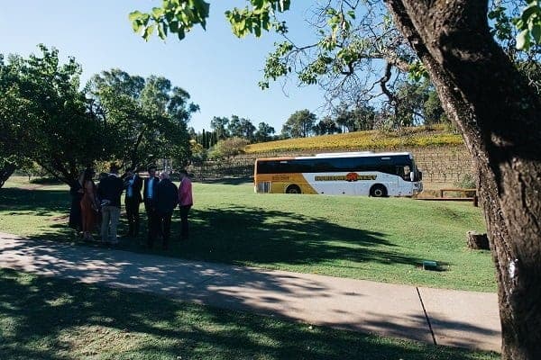 A Horizons West coach parked on a lawn below a planted embankment, with wedding guests gathered in the shade of a tree